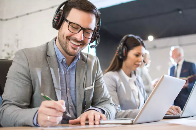 Smiling friendly male customer support agent wearing headset taking some notes while talking to a client in call center